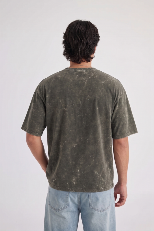 A man viewed from behind wearing a washed olive green t-shirt and light blue jeans, standing against a plain light gray studio background.