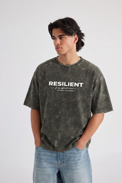 A man wearing a dark olive green acid-wash graphic t-shirt with the word 'RESILIENT' printed in white across the chest, paired with light blue jeans, set against a plain light gray background.