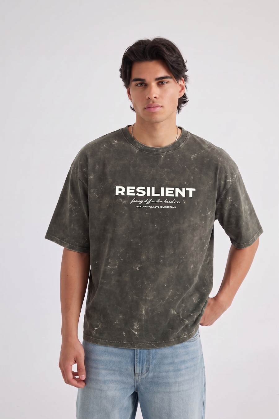 A man wearing a dark gray mineral-wash graphic t-shirt with 'RESILIENT' printed in white across the chest, paired with light blue jeans, standing against a light gray studio background.