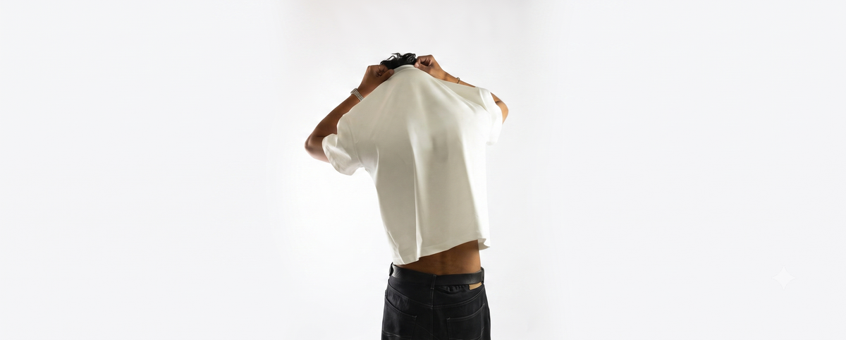 A person wearing a white short-sleeve top and dark jeans, holding the collar of the shirt up to show the back or inner side, against a plain white studio background.