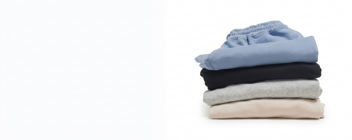 A stack of five folded sweatpants in light blue, black, heather gray, cream, and light beige, photographed on a plain white background.