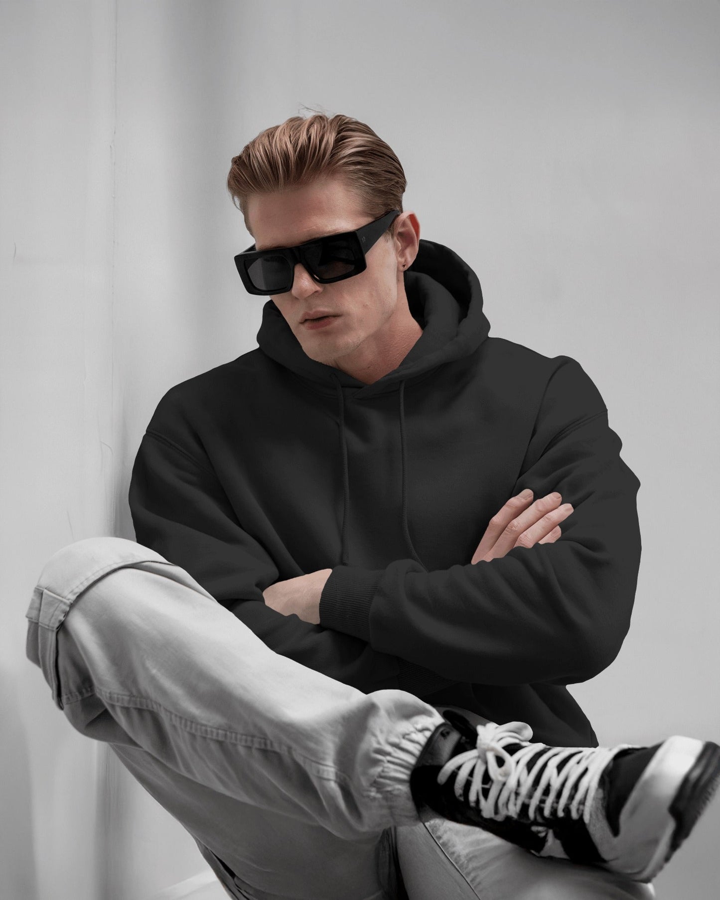 Man wearing a black hoodie, sunglasses, and gray pants sitting against a light gray background