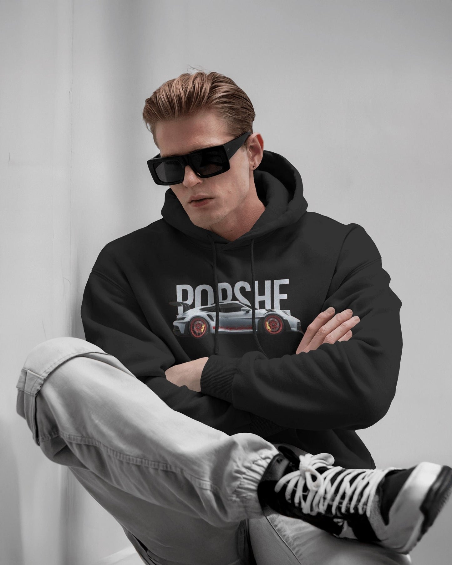 Man wearing a Colour_Black Porsche hoodie with sunglasses and gray pants sitting on a white couch.
